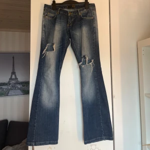 Blå bootcut jeans från Sevan - Midjemått: 35cm  Innerbensmått: 76cm Ytterbensmått: 97cm                                         Säljer ett par blå bootcut jeans från Sevan med slitningar och hål på båda benen framtill. Jeansen har klassisk femficksdesign, låg midja och tydliga slitdetaljer både fram och bak. Perfekta för dig som gillar en avslappnad och trendig look.