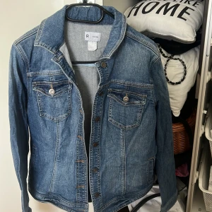 Klassisk blå jeansjacka från Reserved - Snygg blå jeansjacka från Reserved med klassisk krage, bröstfickor med knappar och långa ärmar. Jackan har en figurnära passform och är tillverkad i slitstarkt jeanstyg. Perfekt att slänga på sig över en hoodie eller t-shirt.