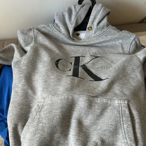 Grå Calvin Klein hoodie med logga - Snygg grå hoodie från Calvin Klein med stor svart CK-logga på bröstet. Tröjan har huva med metallöljett, magficka och ribbade muddar vid ärmslut och nederkant. Perfekt för en chill och stilren look. 