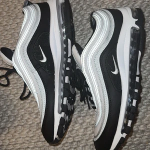 Nike Air Max 97 svart och vit - Nike Air Max 97 sneakers i svart och vitt med ikoniska vågiga linjer och synlig Air-sula. Skorna har snörning, rund tå och reflekterande detaljer. Perfekta för dig som gillar en sportig och stilren look.