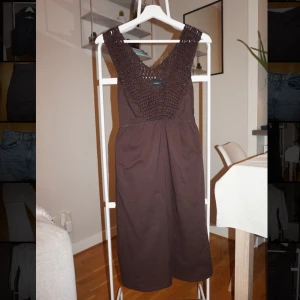 Brun långklänning från Lindex - Snygg brun långklänning från Lindex med virkad överdel och v-ringning. Klänningen har breda axelband, knyt i midjan och är ärmlös. Perfekt för sommarens varma dagar och har en avslappnad men trendig look.