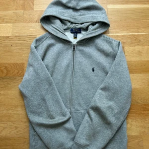 Grå hoodie från Polo Ralph Lauren - Klassisk grå hoodie från Polo Ralph Lauren med dragkedja och broderad marinblå logga på bröstet. Tröjan har huva, långa ärmar och är tillverkad i mjuk bomullsmix. Perfekt för en chill och stilren look.