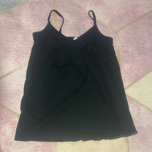 Svart linnetopp med rosett från MyWear - Svart linnetopp från MyWear med smala axelband och v-ringad omlott framtill. Söt liten rosett på ena sidan ger en extra detalj. Perfekt för dig som gillar enkel men ändå söt stil. Tunt och mjukt material som känns skönt mot huden.