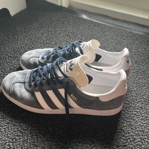 Adidas Gazelle blå/vita sneakers - Säljer ett par klassiska Adidas Gazelle sneakers i blå mocka med vita detaljer och mörkblå snörning. Skorna har de ikoniska tre ränderna på sidan och vit sula. Perfekta för dig som gillar retrostil och vill ha något som sticker ut.