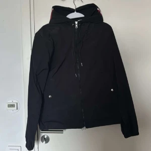 Svart vindjacka från Moncler med huva - Svart vindjacka från Moncler med huva och snygga rödvita detaljer på insidan av huvan. Jackan har dragkedja framtill, två fickor med tryckknappar och Moncler-logga på ärmen. Materialet är glansig polyester och passformen är normal. Perfekt för vår och höst.