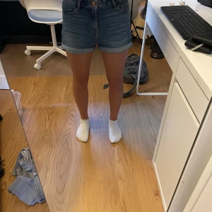 Jeansshorts från Lindex - Snygga blå jeansshorts från Lindex. Shortsen har uppvikta benslut, bälteshällor och stängs med dragkedja och knapp. 