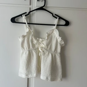 Vit linnetopp med volang från SHEIN - Supersöt vit linnetopp från SHEIN i storlek XS. Toppen har smala axelband, volangdetaljer vid urringningen och en liten knytning framtill. Materialet är lätt skrynklat och ger en chill vibe, perfekt för varma dagar.