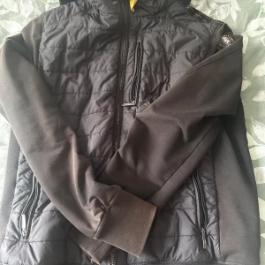 Svart hybridjacka från Parajumpers - Svart hybridjacka från Parajumpers med quiltad front och rygg i mjukare material. Jackan har huva, dragkedjor på fickor och bröstficka samt en snygg patch på ärmen. Perfekt för dig som vill ha en sportig och stilren look.