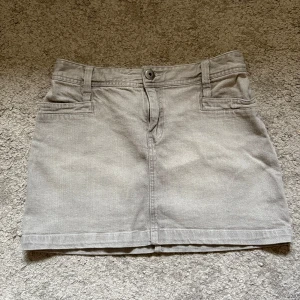 Lowwaist Jeanskjol - Snygg lowwaist grå jeanskjol bältdetalj bak. Kjolen är kort och har hällor för bälte, knapp och dragkedja framtill. 