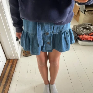 Blå jeanskjol med volang och knappar från Monsoon - Supersöt blå jeanskjol med volangkant och knappar framtill. Kjolen har ett knytband i midjan och en lätt utställd form som ger en chill vibe. ( resår baktill )Perfekt att styla med sneakers eller boots för en avslappnad look.