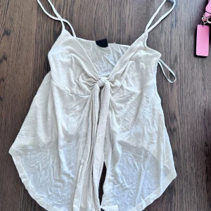 Beige linnetopp med knyt fram från gina - Snygg beige linnetopp med tunna axelband och knytning framtill. Toppen har en luftig passform och är perfekt för varma dagar. Mjuk och lätt i materialet, med en enkel men trendig look.