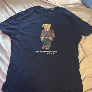 Mörkblå Polo Bear t-shirt Ralph Lauren - Snygg mörkblå t-shirt från Polo Ralph Lauren med ikoniska Polo Bear-trycket på bröstet. Klassisk passform och rund halsringning. Tillverkad i mjuk bomull, perfekt för en chill och stilren look. Bra skick riktigt skön nypris ca 1000