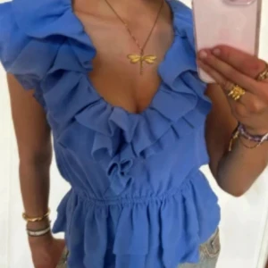 Blå volangblus med djup v-ringning - Säljer denna trendiga supersöt blå blus med volanger längs v-ringningen och axlarna. I väldigt bra skick och tillkommer även ett svart band man kan knyta runt midjan om man skulle vilja❤️ 