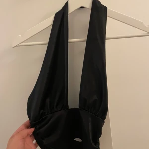 Svart halterneck topp från NLY One - Snygg svart halterneck topp från NLY One i glansigt material. Toppen har djup urringning, breda axelband och stängs med knapp i nacken samt dragkedja i sidan. Perfekt för dig som vill sticka ut med en trendig och stilren look.