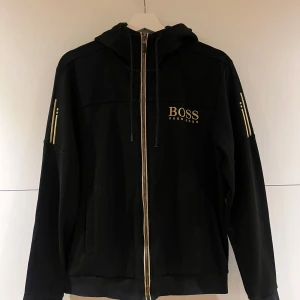 Svart hoodiejacka från Hugo Boss - Svart hoodiejacka från Hugo Boss med guldfärgade detaljer och dragkedja. Jackan har huva med snörning, ribbade muddar och guldiga logotyper på bröstet och ärmarna. Perfekt för en stilren och sportig look.