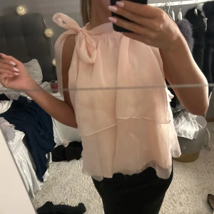 Volang blus - Supersöt ljusrosa blus med volanger och stor rosett på axeln. Ärmlös modell i ett tunt, flowy material som ger en romantisk vibe. Perfekt för dig som gillar en feminin och trendig look.