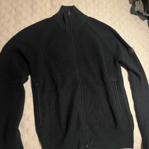 Svart stickad kofta från j.Lindeberg - Svart stickad kofta från j.Lindeberg med hel dragkedja framtill och höga ribbade muddar. Tröjan har två fickor med dragkedja och en stilren, hög krage. Perfekt för lager-på-lager och snygg till både jeans och byxor.