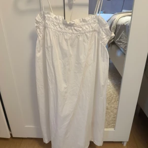 Vit sommarklänning med smock - Söt vit klänning med smala axelband och smockad överdel. Lätt och luftig modell som faller rakt ner, perfekt för varma dagar. Klänningen har en enkel design med volangkant upptill och är tillverkad i ett tunt bomullstyg.