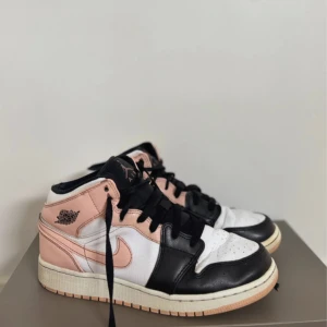 Nike Air Jordan 1 Mid rosa/svart/vit - Nike Air Jordan 1 Mid sneakers i en snygg mix av rosa, svart och vitt skinn. Klassisk Jordan-logga på sidan och svart snörning. Mellanhöga med vit sula och rosa detaljer, perfekt för dig som vill sticka ut med en ikonisk streetstyle.