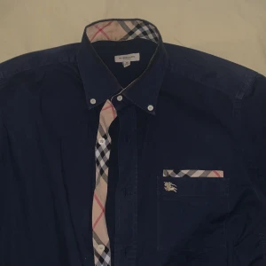 Mörkblå Burberry skjorta med rutiga detaljer - Snygg mörkblå skjorta från Burberry med klassiska rutiga detaljer i beige, svart och rött på knappslå, armbågar, bröstficka och ärmslut. Skjortan har lång ärm, knappar framtill och ikonisk Burberry-logga på bröstet. Perfekt för dig som gillar stilrena och exklusiva plagg.