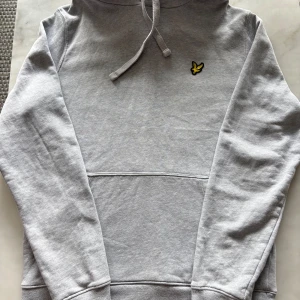 Grå hoodie från Lyle & Scott - Snygg ljusgrå hoodie från Lyle & Scott med klassisk gul logga på bröstet. Tröjan har huva med snörning, magficka och ribbade muddar. Perfekt för en chill och stilren look. Tillverkad i mjuk bomullsmix för extra komfort.