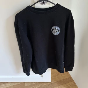 Svart Vans sweatshirt med tryck - Svart sweatshirt från Vans i storlek S med stort rundat tryck bak och mindre logga fram. Klassisk crewneck-modell med långa ärmar och ribbade muddar. Materialet är mjukt och skönt, perfekt för chill dagar. Snygg streetstil med schackrutigt Vans-tryck.