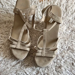 Beige sandaler med kilklack, storlek 38 - Snygga beige sandaler med kilklack och öppen tå. Remmar i mockaimitation och spänne i guld. Sulan har flätad struktur som ger en somrig känsla. Perfekta för dig som gillar stilrena och bekväma skor med lite höjd.