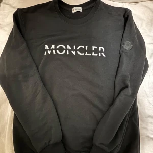 Moncler tröja - Svart Moncler sweatshirt med broderad vit logga på bröstet och en svart Moncler-patch på ärmen. Tröjan har rund halsringning och långa ärmar. Tillverkad i mjuk bomull för en skön och avslappnad stil. Pris kan diskuteras vid snabb affär.