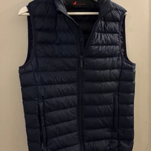 Mörkblå dunväst med dragkedja - Snygg mörkblå dunväst med quiltad design och hel dragkedja framtill. Västen har ståkrage och två sidofickor med dragkedja. Perfekt att slänga över en hoodie för extra värme. Enkel och stilren look som funkar till det mesta.