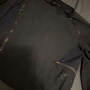 Svart jacka från BOSS med dragkedja - Snygg svart jacka från BOSS med diskreta logodetaljer och dragkedja på sidan. Jackan är lätt, vattenavvisande och har stretchigt material. Perfekt för dig som vill ha en stilren och funktionell jacka med exklusiv känsla.