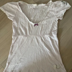 Ljusrosa Odd Molly babydoll  - Söt ljusrosa babydoll top från Odd Molly med en liten lila rosett framtill, i storlek xs. Korta ärmar med spetskant och broderad fjäril på ena ärmen. Köpt second hand men tyvärr passar den inte. Finns ett litet hål och några få lösa trådar