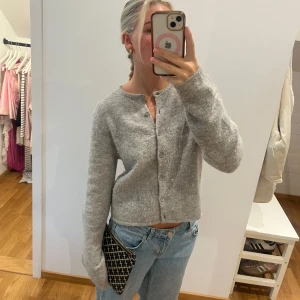 Grå kofta i ull med knappar - Mysig grå kofta i ull med rund hals och knäppning framtill. Modellen är lite croppad och har långa ärmar. Perfekt att slänga över en t-shirt eller bära som den är till jeans.