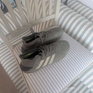 Gråa adidas fotbollsskor med snörning - Snygga grå fotbollsskor från adidas med vita ränder på sidorna och stickad strumpliknande krage. Skorna har snörning och mönstrad ovandel i syntetmaterial. Sulan har fasta dobbar i ljus färg, perfekt för spel på gräs.