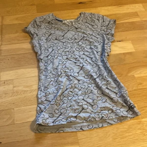 Grå mönstrad topp från Ariat XS - Säljer en grå kortärmad topp från Ariat i storlek XS. Toppen har ett unikt cirkelmönster i mörkare grått och är tillverkad i mjuk bomull. Perfekt för dig som gillar en avslappnad och cool stil.