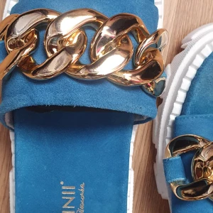 Blå sandaler från Carinii med guldkedja - Snygga blå sandaler från Carinii med chunky vit sula och bred rem i mocka. Dekorerade med en stor guldfärgad kedja över foten som ger en cool statement-look. Perfekta för dig som vill sticka ut med dina skor i sommar.