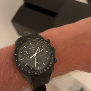 Omega x Swatch Speedmaster MoonSwatch - Grym svart Omega x Swatch Speedmaster MoonSwatch Mission to Mercury med tachymeter och kronograf. Klockan har en rund boett, svart urtavla med vita detaljer och levereras med originalförpackning, extra grått band och verktyg för bandbyte. Perfekt för dig som gillar stil och precision.