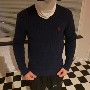 Mörkblå stickad tröja Polo Ralph Lauren - Snygg mörkblå stickad tröja från Polo Ralph Lauren med klassiskt kabelstickat mönster och röd broderad logga på bröstet. Rund hals och långa ärmar, perfekt för dig som gillar preppy stil och vill ha något som funkar till jeans eller kjol.