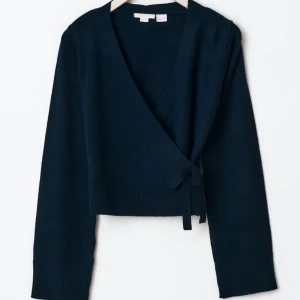 Marinblå omlottkofta med knyt - Snygg marinblå omlottkofta från Wera med knyt i sidan och ribbade muddar. Koftan är croppad och har långärmade ärmar, perfekt att slänga på över en t-shirt eller linne. Mjuk stickad kvalitet som känns skön mot huden.