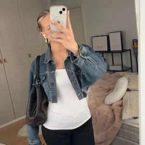 Croppad jeansjacka i blå denim - Säljer en croppad jeansjacka i klassisk blå denim med råa kanter och knappar framtill. Jackan har en loose passform och långa ärmar, perfekt att slänga över en t-shirt eller linne för en chill look.