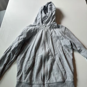 Grå hoodie med dragkedja från Cubus - Grå hoodie från Cubus i storlek S, med hel dragkedja framtill och två stora fickor. Tröjan har huva och är tillverkad i mjuk bomullsmix, perfekt för chill dagar. Klassisk och enkel design som funkar till allt.