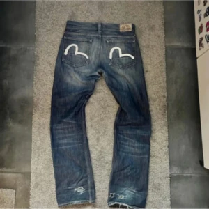 Evisu jeans blå med målad bakficka - Säljer ett par coola blå jeans från Evisu med deras ikoniska handmålade vita måslogga på bakfickorna. Jeansen har en rak passform, bilderna tagna innan de blivit uppsydda, passar ungefär 170-175cm l28. Säljer pga för små för mig.