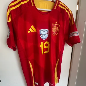 Lamine Yamal Spanien EM 2024 Autentisk Kvalité - Säljer en officiell röd Spanien fotbollströja från EM 2024 med nummer 19. Tröjan har gula detaljer, broderat landslagsemblem, turneringspatchar på ärmarna och Adidas-logga. Tillverkad i lätt och ventilerande funktionsmaterial.