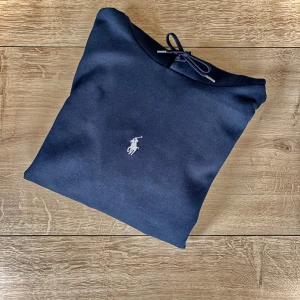 Mörkblå hoodie från Polo Ralph Lauren - Snygg mörkblå hoodie från Polo Ralph Lauren med klassisk vit logga broderad på bröstet. Tröjan har huva med snörning, magficka och ribbade muddar. Perfekt för chill dagar och streetstyle. Materialet är mjuk bomull som känns skönt mot huden.