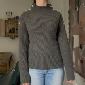 Mörkgrön stickad tröja med guldknappar - Säljer en mörkgrön stickad tröja med ribbad struktur och snygga guldknappar längs axlarna. Tröjan har hög hals och långa ärmar, perfekt för att hålla sig varm och ändå se cool ut. Passar dig som gillar detaljer och vill ha något som sticker ut lite extra.