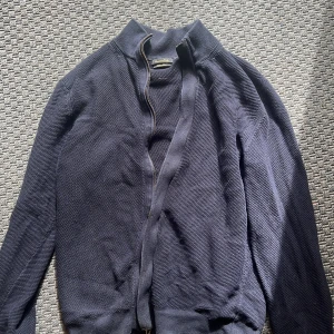 Mörkblå stickad kofta från Massimo Dutti - Säljer en mörkblå stickad kofta från Massimo Dutti med grov struktur och hög krage. Koftan har en snygg dragkedja framtill och ribbade muddar vid hals och ärmslut. Perfekt för dig som gillar stilrena och klassiska plagg med lite edge. Storlek M men sitter lite som M/S. Använd fåtal ggr