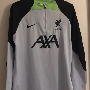 Liverpool tröja   - Säljer en snygg Liverpool FC träningströja från Nike med half zip och långärmad design. Tröjan är ljusgrå med svarta och gröna detaljer, samt stora AXA-loggan och LFC-märket på bröstet. Perfekt för fotbollsträning eller chill.