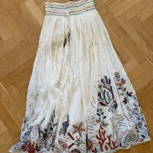 Vit Zara långkjol  - Superfin vit långkjol med färgglada broderier av havsmotiv längst ner och smockad midja med snörning. Kjolen är luftig och har en bohemisk vibe, perfekt för varma dagar. Materialet känns som bomull och midjan har ett vågigt mönster i flera färger.
