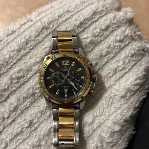 Festina kronograf herrklocka guld/silver - Stilren Festina herrklocka med robust länkarmband i guld och silverfärgat stål. Svart urtavla med guldiga detaljer, tre subdials och datumvisning. Klockan har tydliga index och siffran 12 i guld, samt kronograf-funktion och vridbar ring.