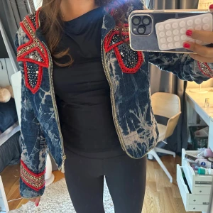 Quiltad jeansjacka från Ambika L - Unik quiltad jeansjacka från Ambika i blått med batikmönster, röda broderade detaljer och pärlor på axlar och ärmslut. Jackan har dragkedja framtill och coola detaljer som verkligen sticker ut. Perfekt för dig som vill ha något annorlunda och färgstarkt. Storlek L men passar mig som vanligtvis har storlek S