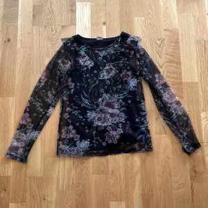 Svart blommig meshblus från Lindex - Säljer en svart meshblus från Lindex med transparenta långärmar och ett snyggt blommigt mönster i rosa, blått och vitt. Perfekt för dig som gillar detaljer och vill sticka ut lite extra. Blusen har en lätt och luftig känsla som passar till många olika looks.den har genomskinliga armar men svart över magen